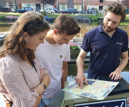 Varen Amsterdamse grachten mooie vaarroutes Boaty Een man legt de vaarroutes in Amsterdam uit aan een man en vrouw.