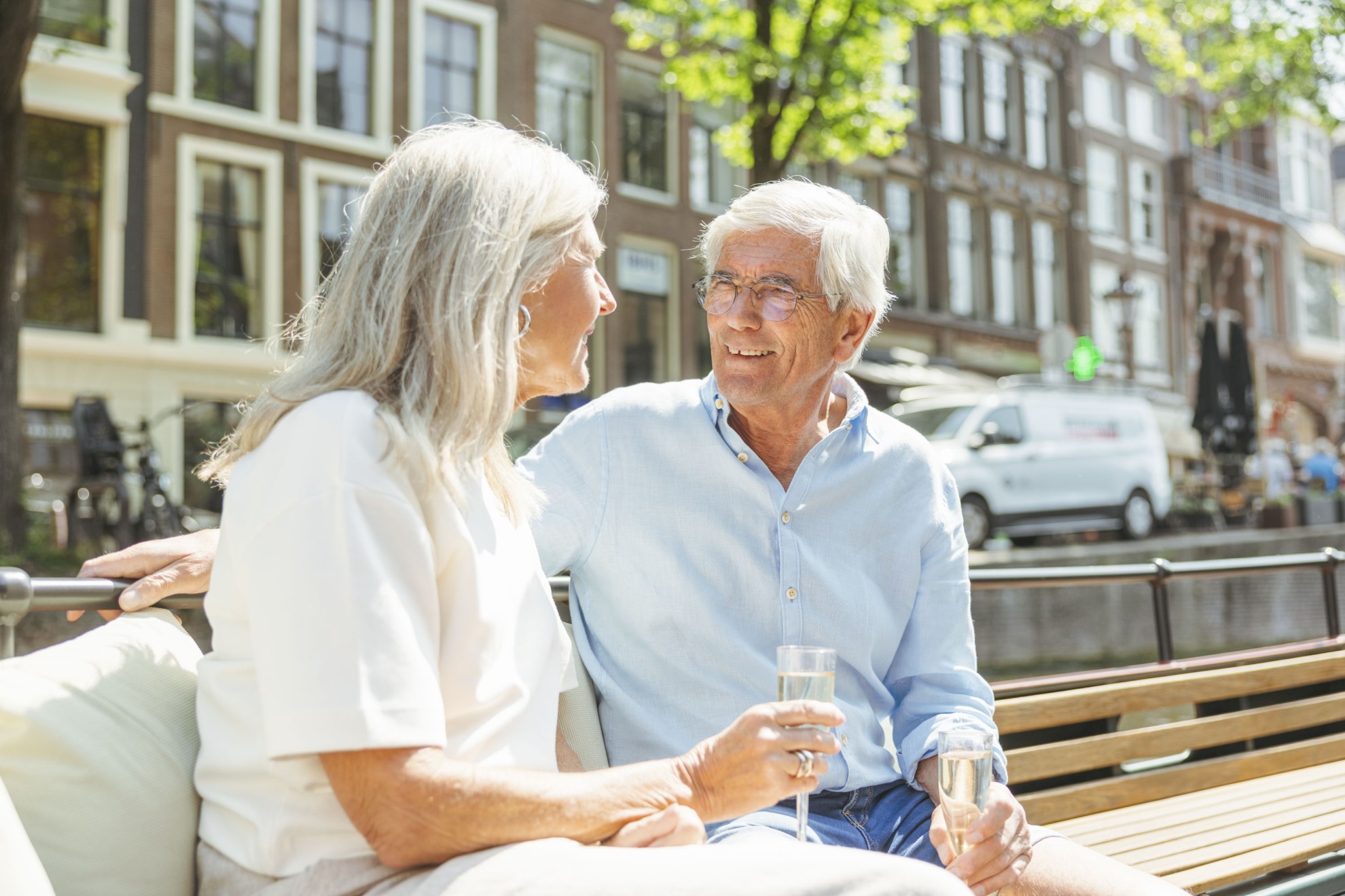 Een ouder stel zit ontspannen op een boot in de Amsterdamse grachten, glimlachend naar elkaar terwijl ze genieten van een glas wijn.