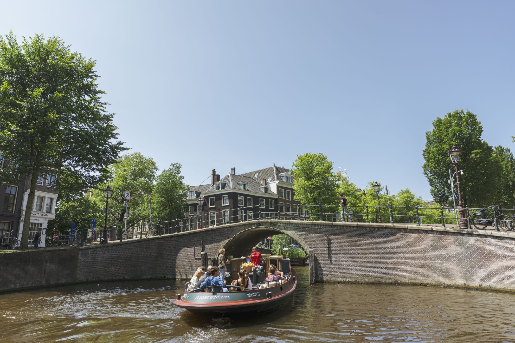 Een boot vol passagiers vaart onder een stenen brug door op de Amsterdamse grachten, omgeven door historische panden en groen.