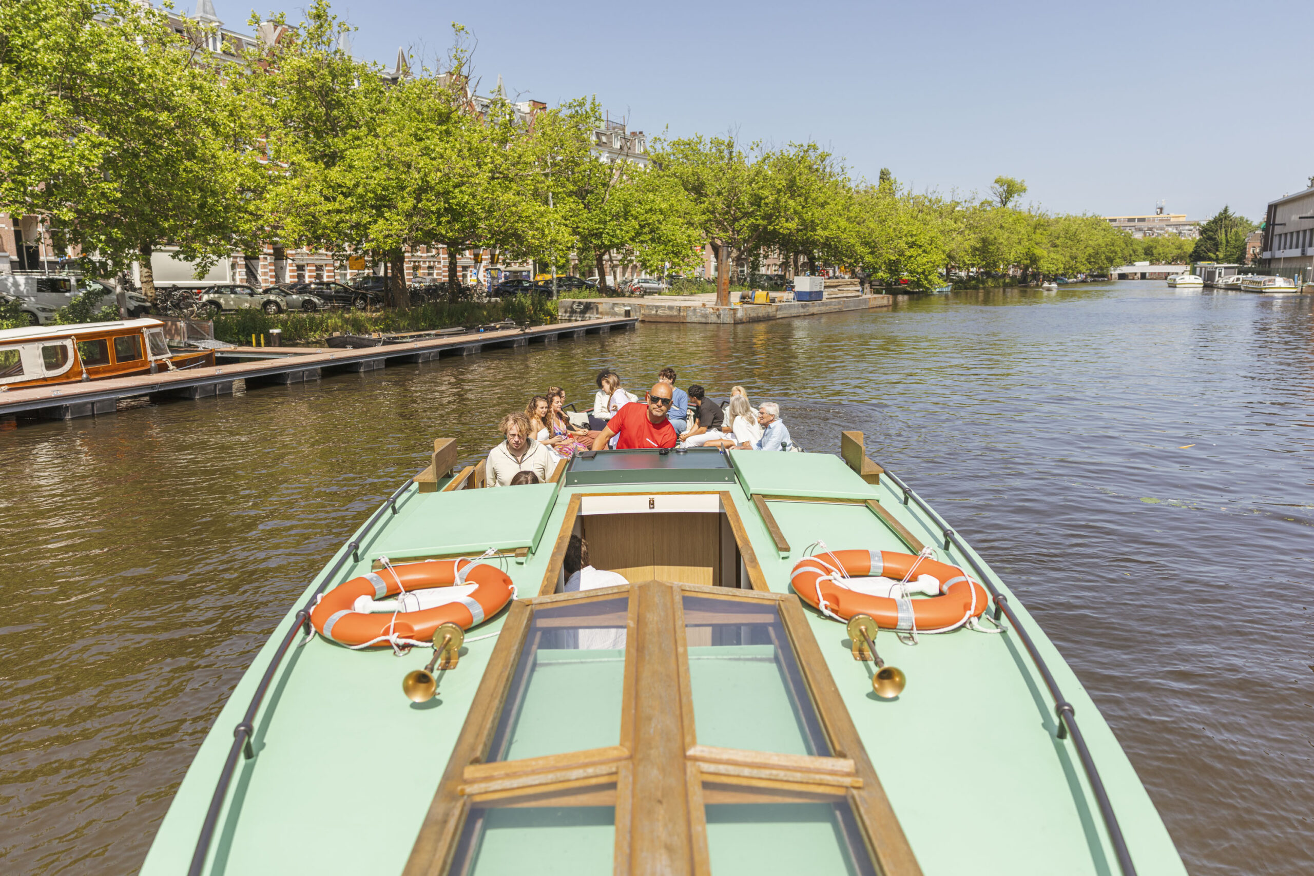 Een groep mensen zit ontspannen op een klassieke rondvaartboot in Amsterdam, terwijl de schipper het vaartuig over de gracht stuurt op een zonnige dag.