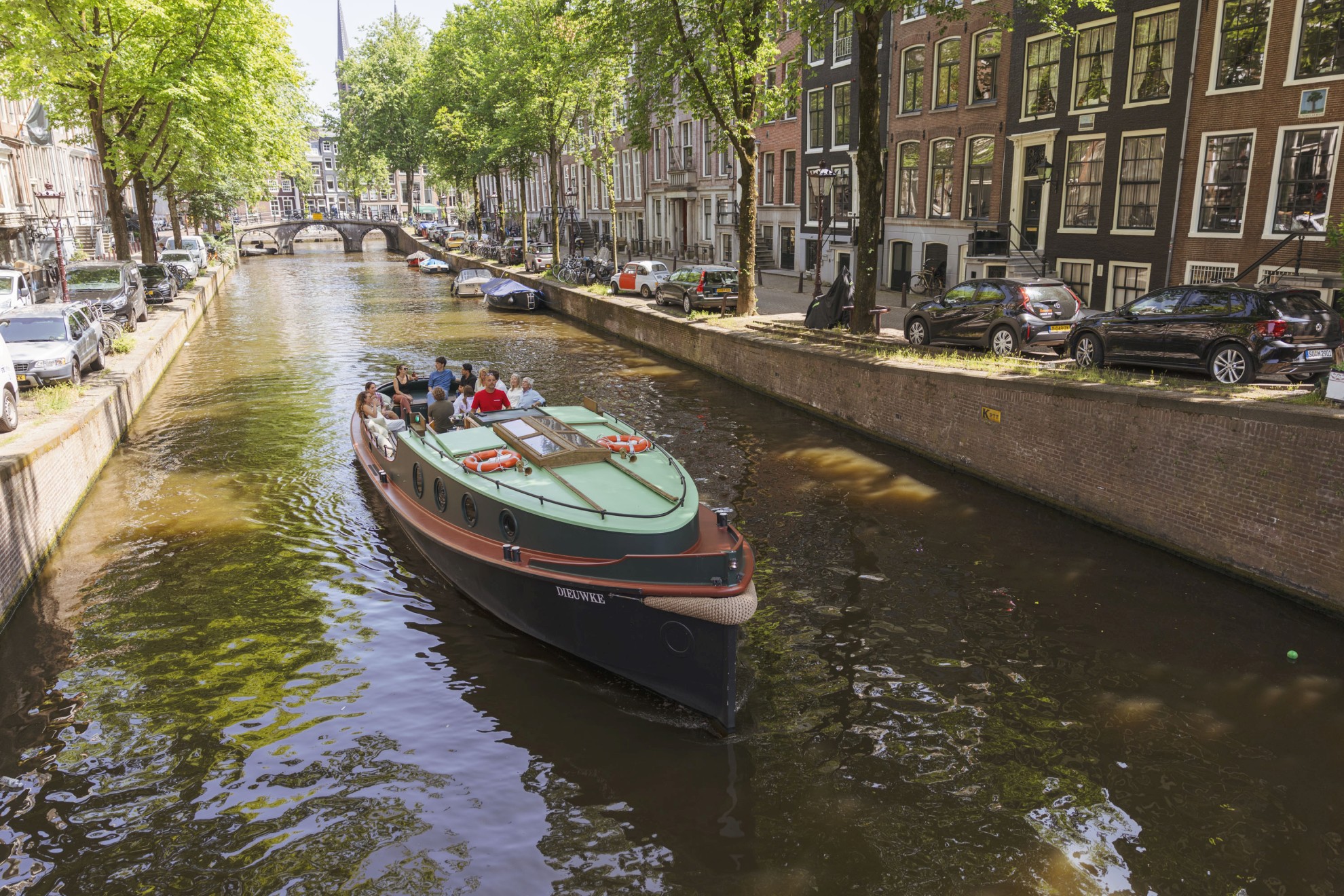 Een groep mensen vaart met een klassieke boot door een Amsterdamse gracht, omgeven door bomen, geparkeerde auto’s en historische grachtenpanden.