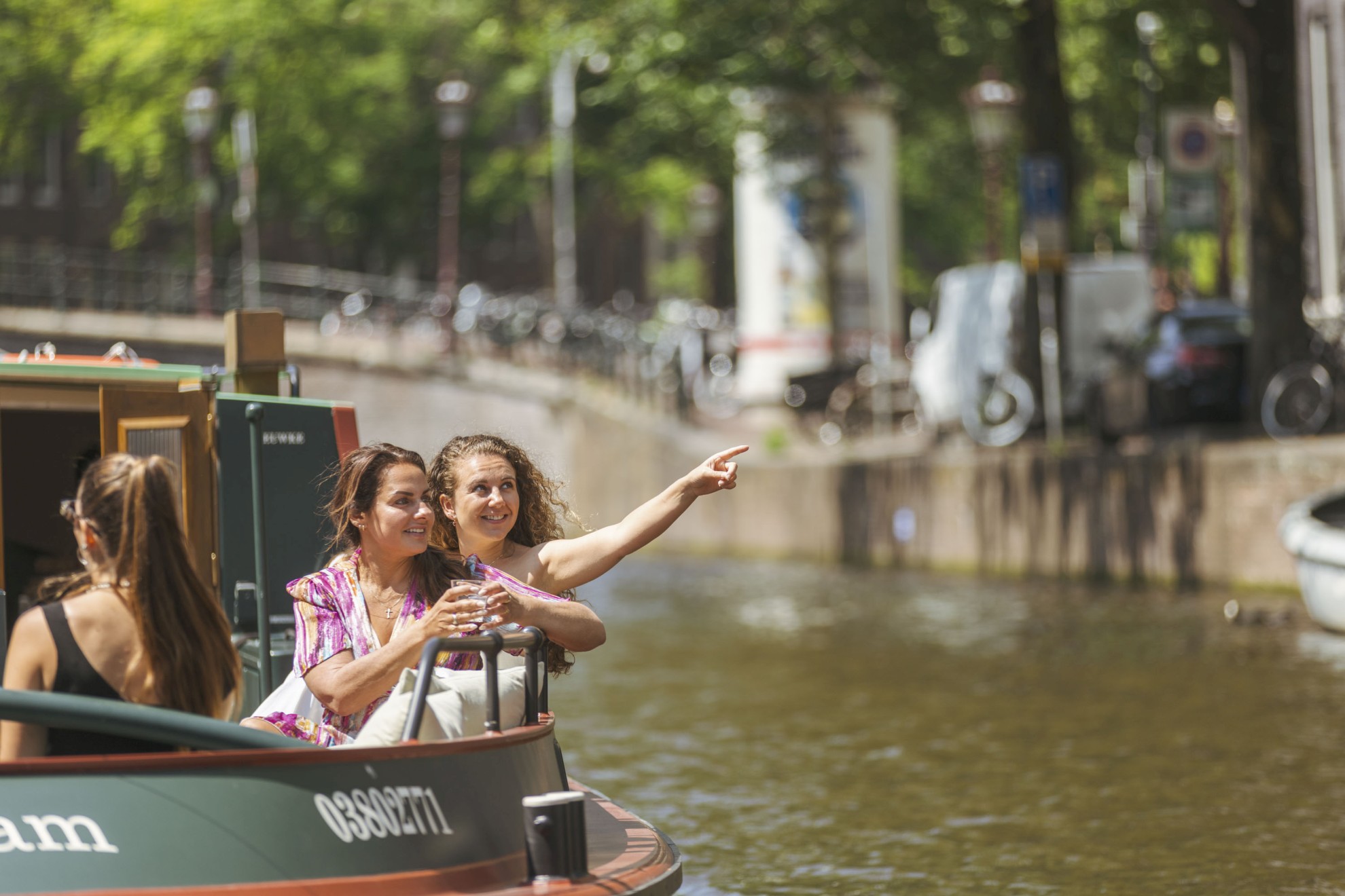 Twee vrouwen genieten van een rondvaart door de Amsterdamse grachten; één van hen wijst enthousiast naar iets in de verte.