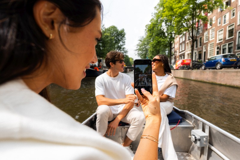 Vrienden maken een foto tijdens een ontspannen boottocht door een Amsterdamse gracht, omringd door historische grachtenpanden en groene bomen.