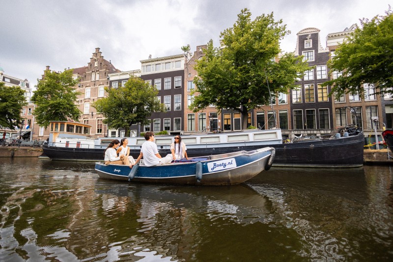 Groep vrienden vaart met een kleine huurboot door een Amsterdamse gracht langs historische grachtenpanden en woonboten.