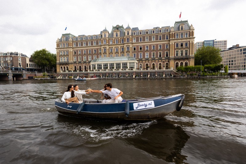 Vrienden proosten met drankjes tijdens een boottocht over het water met op de achtergrond een historisch gebouw langs de Amsterdamse gracht.